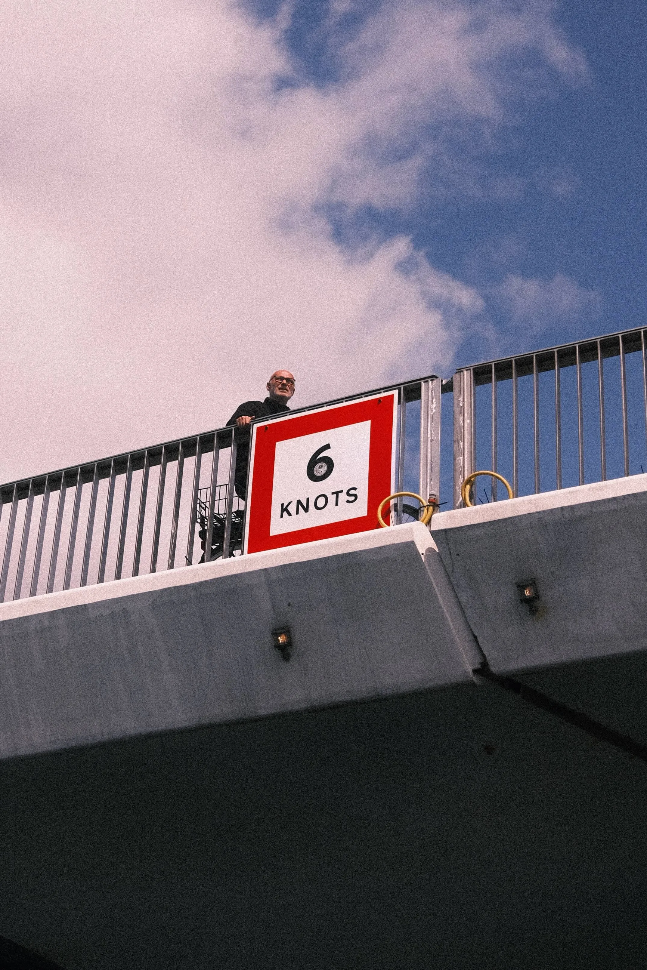 Man standing on bridge above water in copenhagen next to 6 knots sign